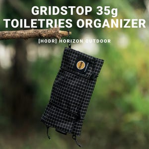 May include: A black and white grid-patterned toiletry organizer, suspended from a branch. The text "GRIDSTOP 35g TOILETRIES ORGANIZER" is displayed above the organizer. The brand name "[HODR] HORIZON OUTDOOR" is also visible.