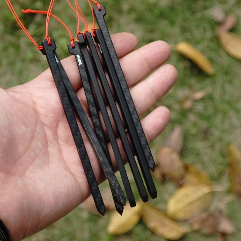 May include: A set of four black carbon fiber tent stakes with a visible woven pattern. Each stake features a pointed tip at the bottom and a notched top attached to a thin, bright orange cord. The brand name HODR is printed in white on the stakes.