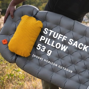 Könnte beinhalten: Eine graue aufblasbare Schlafmatte mit einem gelben Kissen und einem orangefarbenen Ventil. Das Kissen besteht aus einem strukturierten Material. Der Text auf der Matte lautet "STUFF SACK PILLOW 53 g" und "[HODR] HORIZON OUTDOOR".