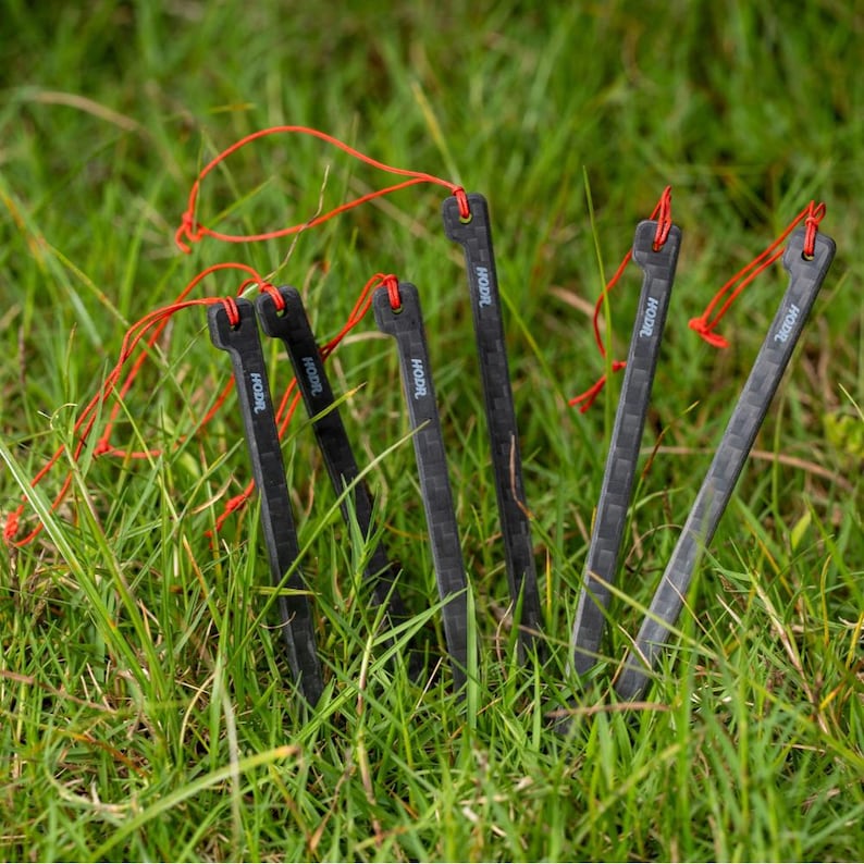 May include: A set of six black tent stakes with a carbon fiber texture, each featuring a small hole at the top and a thin red cord loop. The word HODR is printed in white on the upper section of each stake, which are arranged upright.
