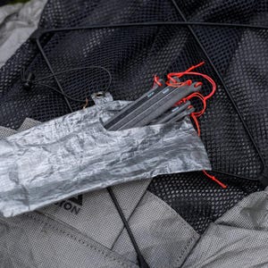 May include: A set of dark gray tent stakes with bright red pull cords, partially inserted into a crinkled, translucent silver storage pouch. The stakes are made of a rigid material and feature a flat, elongated design with a small hole at the top.