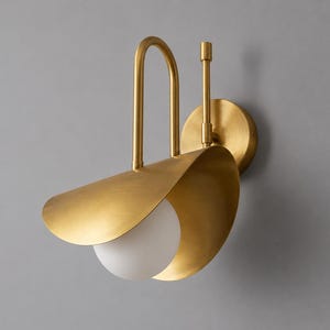 Peut inclure: Une applique murale en laiton brossé dotée d'un abat-jour métallique incurvé en forme de pétale et d'une ampoule sphérique en verre blanc. Le luminaire comprend un bras vertical arqué et une plaque de fixation murale circulaire en laiton.
