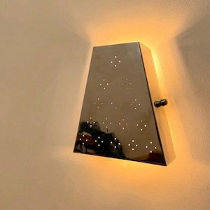May include: A modern, metallic wall sconce with a trapezoidal shape. The sconce has a series of small, evenly spaced holes that emit light. A small, black knob is on the side. The sconce is mounted on a light-colored wall.