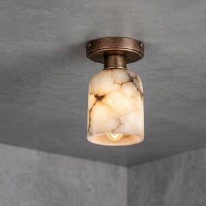 Könnte beinhalten: Eine Deckenleuchte mit einem zylindrischen Schirm aus cremefarbenem Stein mit dunkelbrauner Maserung. Die Leuchte verfügt über einen runden, dunkelbronzefarbenen Metallsockel, der direkt an der Deckenoberfläche befestigt ist.