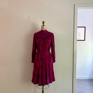 May include: A vintage, long-sleeved dress with a high neck and a floral print in shades of pink and blue. The dress has a fitted bodice and a flowing skirt with a ruffled hem. The dress is trimmed with a blue ribbon.