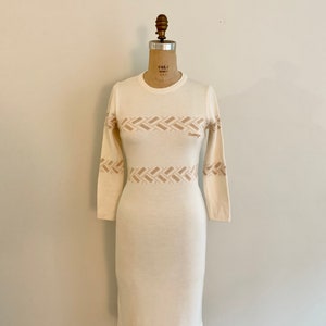 May include: A white long-sleeved sweater dress with a brown geometric pattern around the waist. The dress has a crew neck and is fitted.