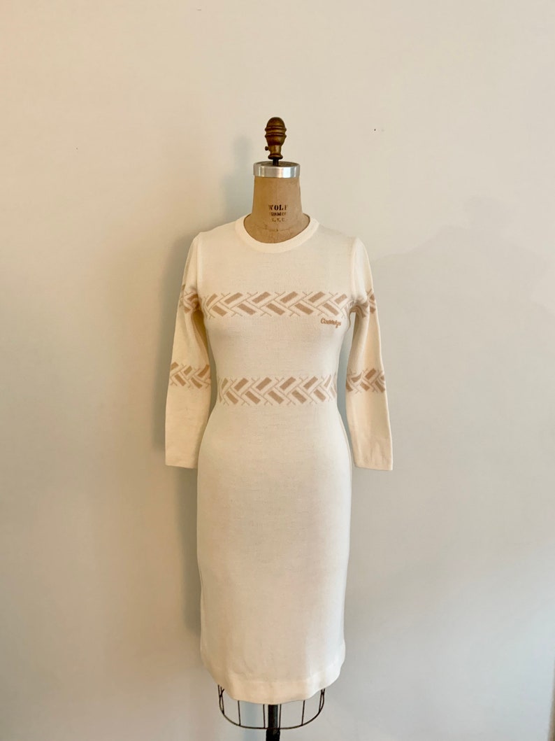 May include: A white long-sleeved sweater dress with a brown geometric pattern around the waist. The dress has a crew neck and is fitted.