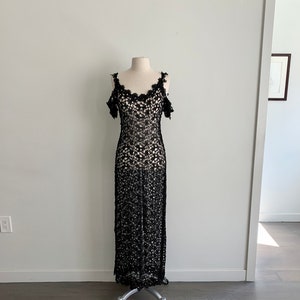 May include: A black, long, crochet dress with a scoop neck and cold shoulder straps. The dress is on a white mannequin against a white wall.