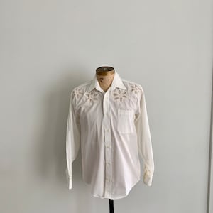 Puede incluir: Una camisa blanca de manga larga con detalles florales recortados en los hombros y el pecho. La camisa tiene una botonadura frontal, un bolsillo en el pecho y un cuello clásico. La camisa se exhibe en un maniquí.