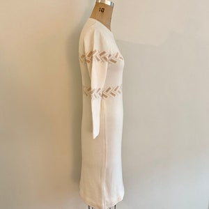 May include: A white knitted dress with a brown woven pattern around the waist. The dress is on a mannequin.