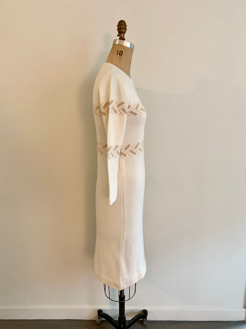 May include: A white knitted dress with a brown woven pattern around the waist. The dress is on a mannequin.