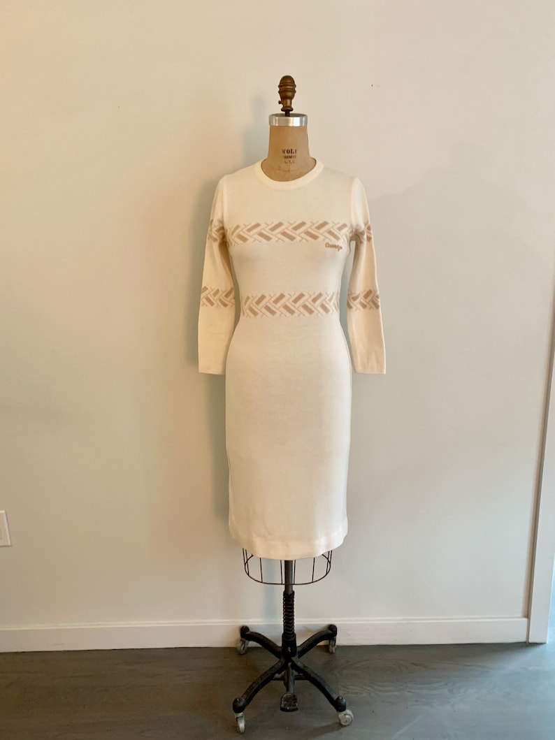 May include: A white long-sleeved sweater dress with a brown geometric pattern around the waist. The dress is on a white mannequin.