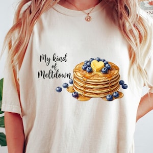Può includere: Una t-shirt color crema con la grafica di una pila di pancake guarnita con un pezzetto di burro a forma di cuore e mirtilli freschi, con il testo "My kind of Meltdown" stampato in caratteri corsivi neri a sinistra della pila.