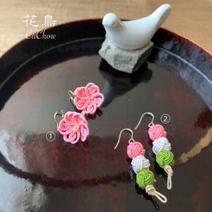 May include: Pink flower-shaped earrings and dangling earrings with pink, white, and green knotted designs. A white bird figurine sits on a gray stone. The image includes the text "花鳥 CaChow".