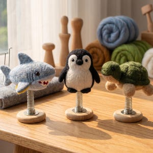 May include: Three needle-felted animal figures on metal springs and wooden bases: a light blue shark with white teeth, a black and white penguin, and a green and tan turtle. Each figure is crafted from wool and mounted on a silver-colored coil spring.