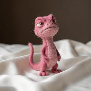 May include: A needle-felted pink lizard figurine with a textured, fuzzy surface. The creature stands upright on two legs with a long, curved tail, small arms, and a distinct facial expression featuring heavy eyelids and a downturned mouth.