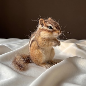 Könnte beinhalten: Eine nadelgefilzte Streifenhörnchen-Figur aus texturierten braunen, beigen und cremefarbenen Wollfasern. Das kleine Tier ist in sitzender Position dargestellt, mit einem buschigen Schwanz, dunklen Augen und markanten Streifen auf dem Rücken.