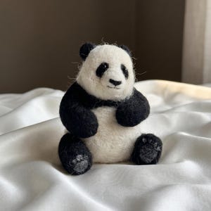 May include: A small, handcrafted needle-felted panda figurine featuring a white face and belly with black ears, limbs, and eye patches. The panda is depicted in a seated position with visible textured wool fibers and small grey paw pads on its feet.