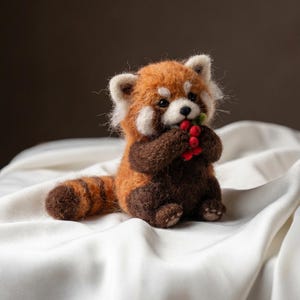 May include: A needle-felted red panda figurine with a fuzzy orange and brown coat, white facial markings, and black eyes. The small creature is sitting upright and holding a cluster of tiny red berries with green stems in its front paws.