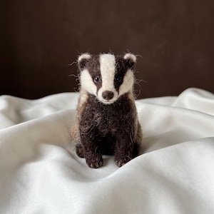 May include: A needle-felted badger figurine featuring a textured dark brown and white coat. The badger has a distinct white stripe running from its forehead to its nose, small rounded ears, dark eyes, and a small brown nose, sitting in an upright position.