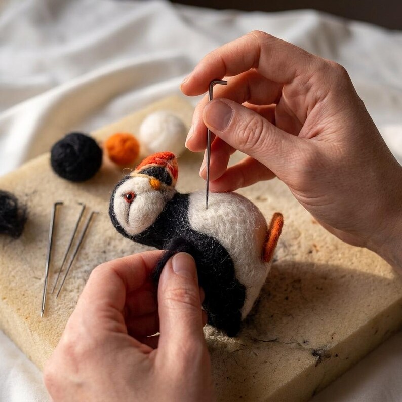 May include: A needle felting project featuring a small puffin figurine made of white, black, and orange wool. The puffin has a distinct orange beak and is being worked on with a metal felting needle. Nearby are loose wool balls and needles.