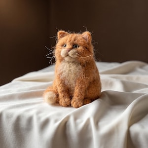 May include: A needle-felted orange tabby cat figurine with a textured, fuzzy coat, a lighter cream-coloured chest, small pointed ears, dark eyes, and long white whiskers. The cat is sitting in an upright position with its front paws together.