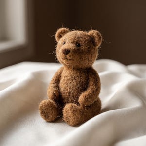 Peut inclure: Un petit ours en peluche fabriqué à la main en fibres de laine brune texturées. L'ours présente une tête arrondie avec de petites oreilles, un nez cousu et des yeux sombres en forme de perles. Il est représenté assis, bras et jambes vers l'avant.