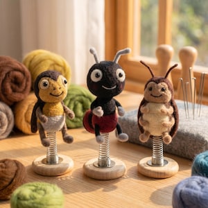 May include: Three handmade felt insect figurines: a beetle, an ant, and a ladybug, each mounted on a wooden base with a spring. The ant is black with a red abdomen, the beetle is brown and tan, and the ladybug is brown and tan. Colorful wool balls are in the background.