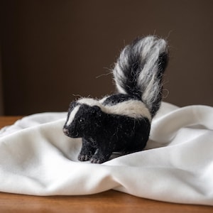 May include: A needle-felted skunk figurine featuring a black body with a white stripe running from the top of its head down its back. The skunk has a large, upright, fluffy tail with black and white fibres and small, dark, bead-like eyes.