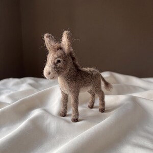May include: A needle-felted donkey figurine crafted from textured, light brown and grey wool fibres. The small animal sculpture features upright ears, a short mane, a thin tail, and a light-coloured muzzle, standing on four slender legs.