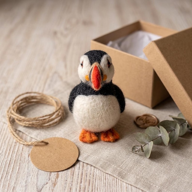 May include: A needle-felted puffin figurine with a black and white body, a bright orange beak, and orange feet. The puffin stands upright, featuring a rounded shape and textured wool fibers, presented alongside a brown cardboard gift box.