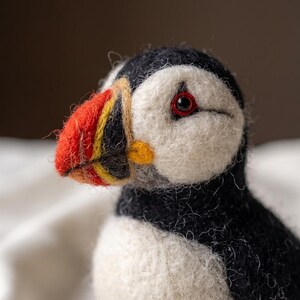 May include: A needle-felted puffin figurine featuring a black and white body, a white face, and a multi-colored beak with vibrant orange, yellow, and black sections. The bird has a single visible dark eye with a red rim and a textured wool finish.