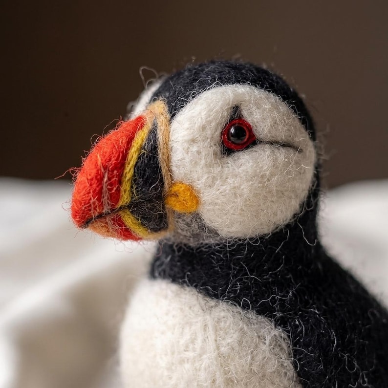 May include: A needle-felted puffin figurine featuring a black and white body, a white face, and a multi-colored beak with vibrant orange, yellow, and black sections. The bird has a single visible dark eye with a red rim and a textured wool finish.