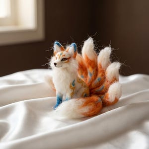 Puede incluir: Una figura de zorro de lana fieltrada con nueve colas esponjosas, con pelaje blanco, naranja y azul. El zorro presenta intrincados patrones de remolinos azules y dorados en el cuerpo, orejas puntiagudas con puntas azules y textura suave.