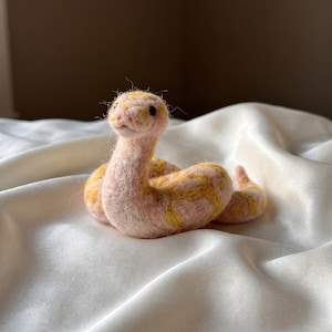 May include: A needle-felted snake figurine crafted from soft fibres in a pale pink hue with distinct mustard yellow patches along its coiled body. The snake features a rounded head, small dark eyes, and a textured, fuzzy surface throughout its form.