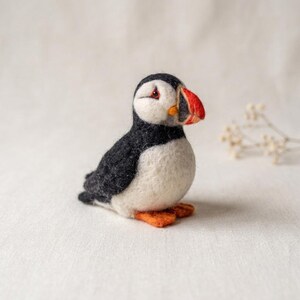 May include: A needle-felted puffin figurine featuring a black back and wings, a white chest and face, and a distinctive beak colored in shades of red, orange, and black. The small bird has orange feet and is crafted with a soft, textured wool finish.