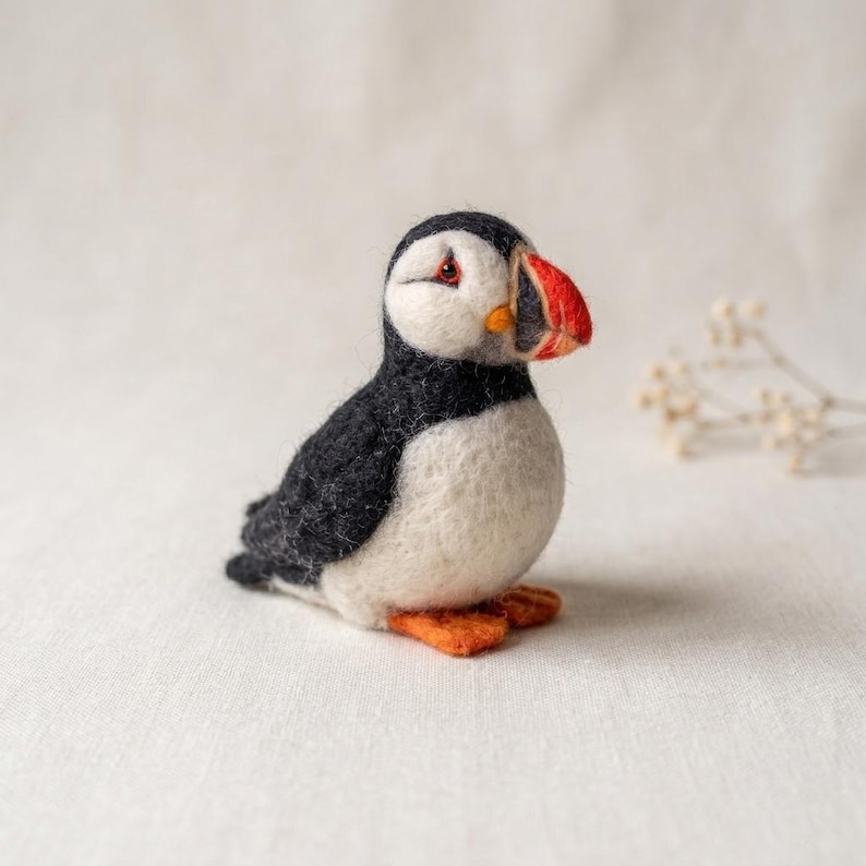 May include: A needle-felted puffin figurine featuring a black back and wings, a white chest and face, and a distinctive beak colored in shades of red, orange, and black. The small bird has orange feet and is crafted with a soft, textured wool finish.