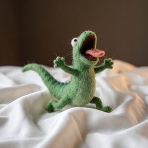 May include: A needle-felted green lizard figurine with a textured wool surface, featuring a wide-open mouth with a pink tongue, large white eyes with black pupils, raised arms, and a long tail, standing in a dynamic, upright, and expressive pose.