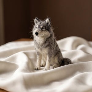 May include: A needle-felted wolf figurine crafted from textured wool fibers in shades of gray, white, and charcoal. The wolf is depicted in a seated position with pointed ears, a black nose, and detailed facial features, showcasing a soft, fuzzy appearance.