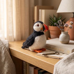 May include: A needle-felted puffin figurine with a black back, white chest, and a distinctive orange, black, and yellow beak. The puffin is positioned on a wooden surface next to an open book and a small ceramic vase containing dried lavender.