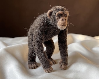 Op de afbeelding: Een naaldgevilte chimpansee-figuur gemaakt van donkerbruine en antracietkleurige wolvezels. Het figuur heeft een getextureerde, pluizige vacht, een lichtbruin gezicht met gedetailleerde donkere ogen en een realistische houding met lange armen.