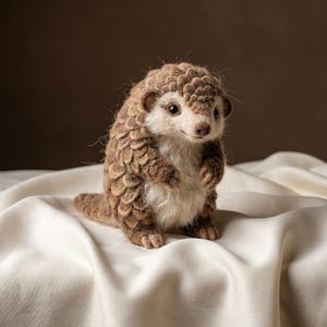 Peut inclure: Une petite figurine de pangolin en feutre faite à la main, présentant un motif d'écailles texturées et superposées dans des tons marron. La créature a un ventre crème, des yeux sombres et un petit museau, debout avec des pattes avant visibles.