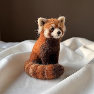 May include: A needle felted red panda figurine featuring a textured coat of rust-orange and dark brown wool fibers. The small animal has white facial markings around the eyes and muzzle, pointed ears, and a curled tail resting on the base.