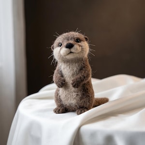 Könnte beinhalten: Eine nadelgefilzte Otterfigur mit einem strukturierten braunen und cremefarbenen Fell, kleinen dunklen Augen, einer schwarzen Nase und ausgeprägten Schnurrhaaren. Der Otter steht aufrecht in einer neugierigen Pose und zeigt detaillierte Handwerkskunst.