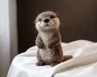 Op de afbeelding: Een naaldgevilte otterfiguur met een textuurrijke bruine en crèmekleurige vacht, kleine donkere ogen, een zwarte neus en opvallende snorharen. De otter staat rechtop in een nieuwsgierige houding en toont gedetailleerd handwerk en zachte vezels.