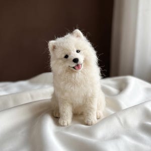 May include: A needle-felted sculpture of a fluffy white Samoyed dog sitting upright. The figure features a textured, wool-like coat, small pointed ears, dark eyes, a black nose, and an open mouth showing a small pink tongue.