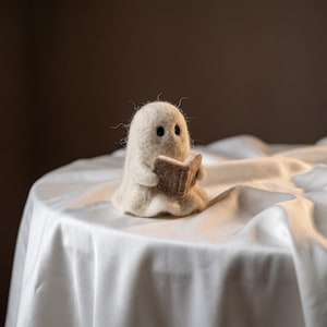 Puede incluir: Una pequeña figura de fantasma de fieltro de aguja en color blanco roto con dos ojos negros redondos. El fantasma sostiene un pequeño libro rectangular de color marrón claro en sus manos, como si estuviera leyendo. La textura es suave y fibrosa.