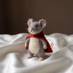 May include: A needle-felted mouse figurine with grey fur, a cream-coloured belly, and small pink paws. The mouse is wearing a deep red cape that drapes over its shoulders and back. It has dark eyes, a small pink nose, and rounded ears.