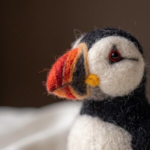 May include: A needle-felted puffin figurine featuring a textured black and white wool body, a bright orange, yellow, and black beak, and a small dark eye with a red ring. The craftwork displays visible wool fibers and a soft, rounded shape.