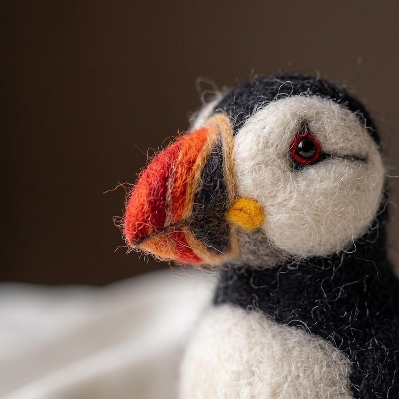 May include: A needle-felted puffin figurine featuring a textured black and white wool body, a bright orange, yellow, and black beak, and a small dark eye with a red ring. The craftwork displays visible wool fibers and a soft, rounded shape.